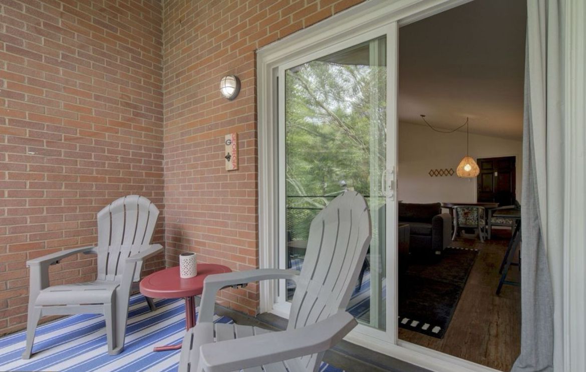 A balcony with two chairs and a small table, a brick wall, and a sliding glass door leading to an interior space with a hanging light fixture.