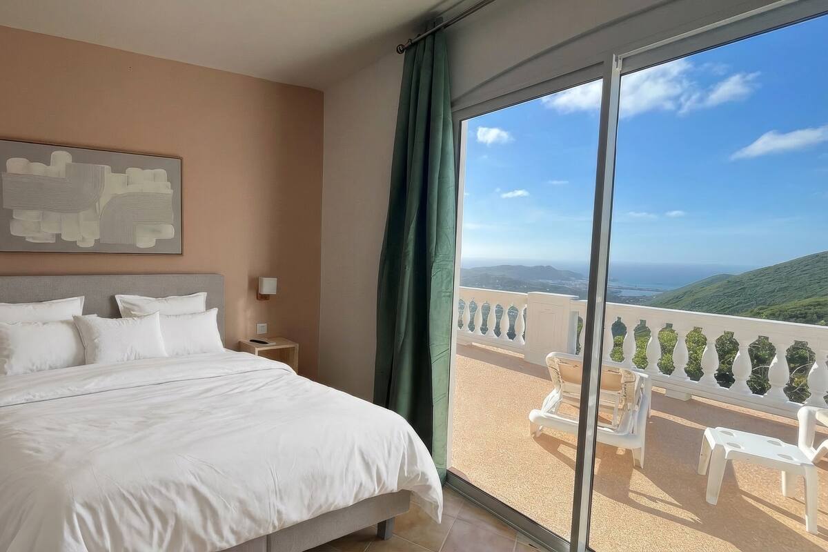 Een hotelkamer met een bed, een schilderij aan de muur, een nachtkastje, een raam met uitzicht op zee en heuvels, en een balkon met stoelen.