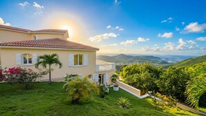 A two-story house with a red-tiled roof, white shutters, and a balcony. The property features a lush green lawn with various plants and trees. - The Pearl of Paradise (Marigot)