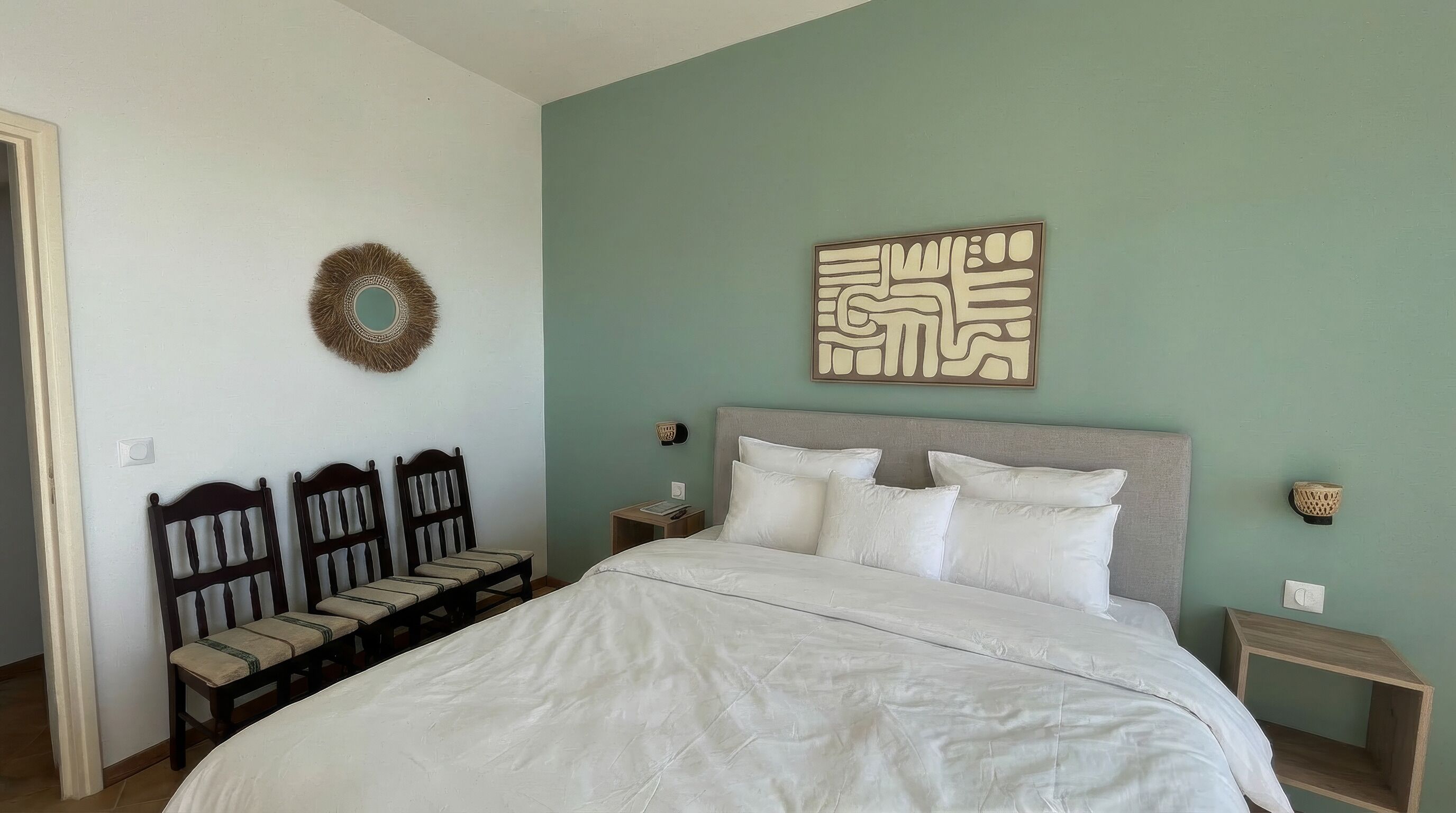 Un dormitorio con cama, cabecera, mesita de noche, una obra de arte en la pared y una corona decorativa.