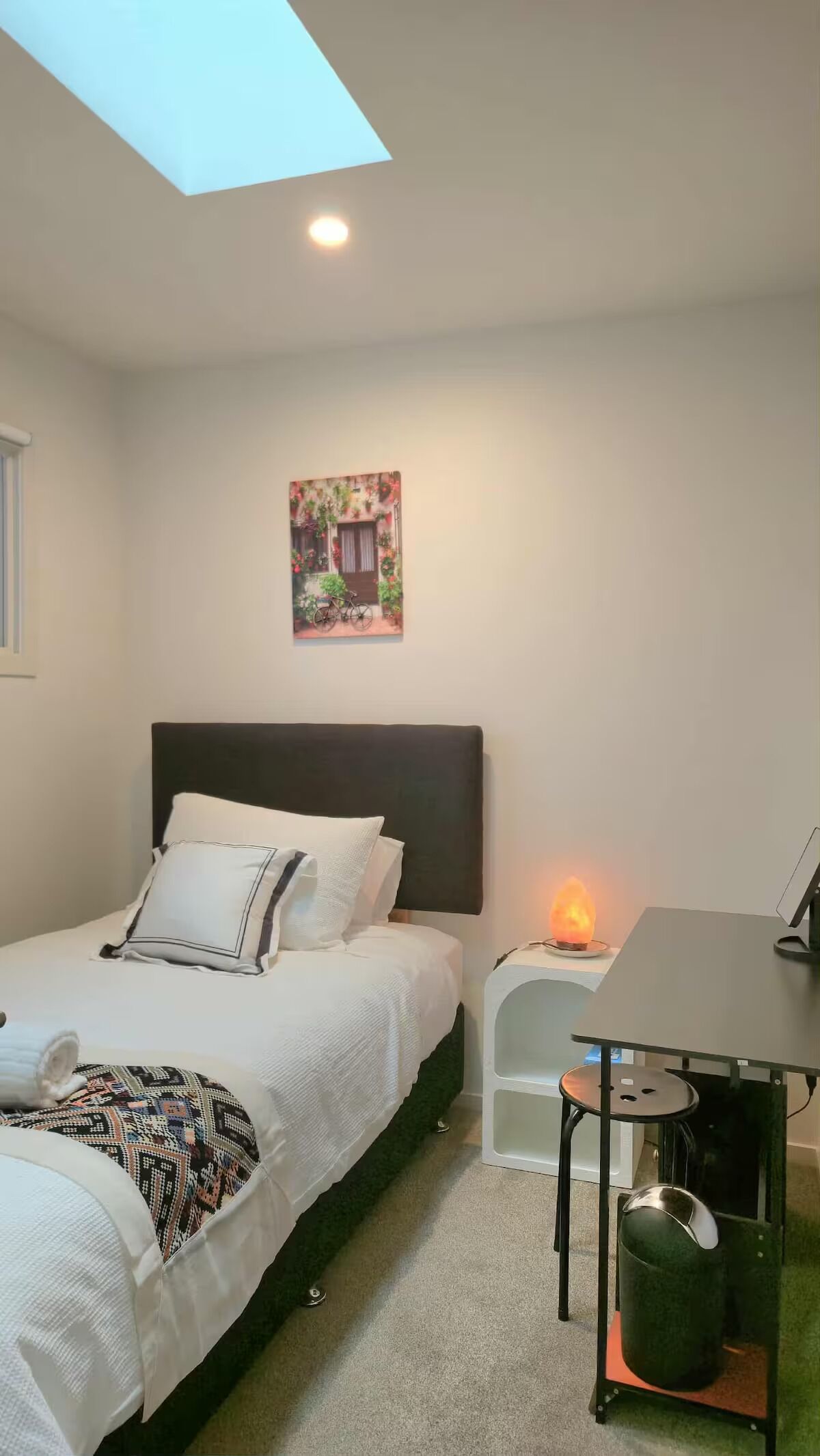 Un dormitorio con cama, escritorio, silla, cubo de basura, calentador en la pared y tragaluz.