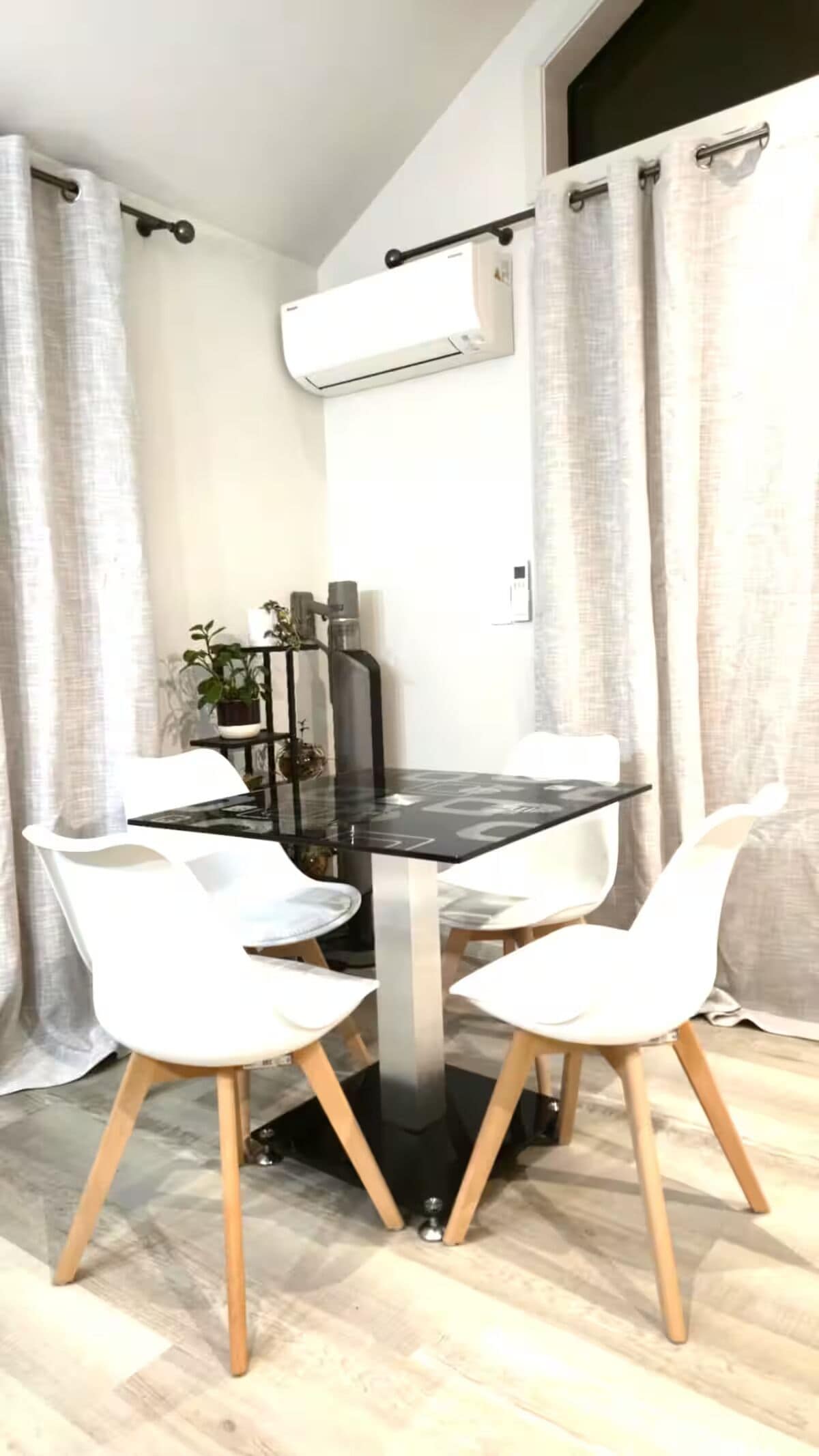 Een eetruimte met een zwarte tafel, vier witte stoelen, een potplant en een airconditioning op de muur.