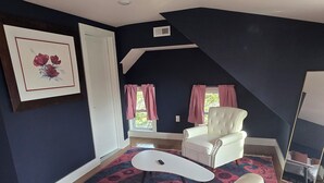 Ein Zimmer mit einem weißen Ledersofa, einem weißen Sessel und einem kleinen weißen Tisch. Auf dem Boden liegt ein farbenfroher Teppich, und an der Wand hängt ein gerahmtes Bild.
