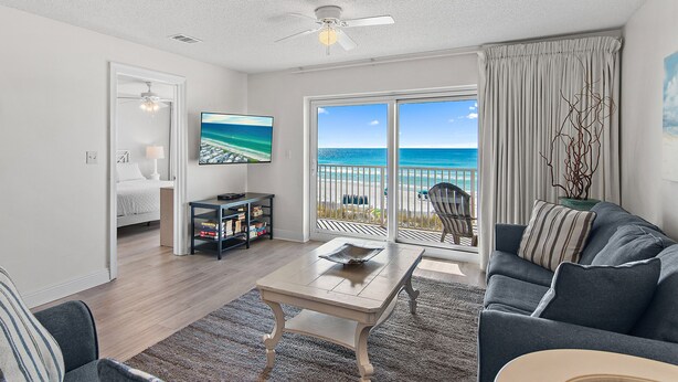 A living room with a large window, a ceiling fan, a flat-screen TV, and a view of the beach. - Emerald Dunes 204*Pool*HotTub*Remodeled*Beach Gear (Destin)