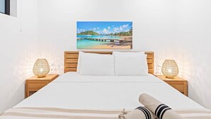 Quarto com cama, cabeceira, mesa de cabeceira, luminária e pintura de uma praia.