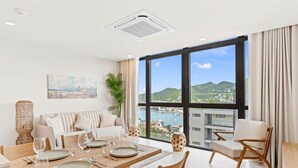 Uma sala de estar moderna com área de jantar, grandes janelas com vista para o mar e colinas, e um ambiente acolhedor para relaxar.