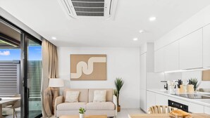 Ein modernes Wohnzimmer mit Sofa, Couchtisch und Esstisch. Es gibt eine Küchenzeile mit Schränken und einer Arbeitsplatte. Ein großes Fenster bietet einen Blick ins Freie.
