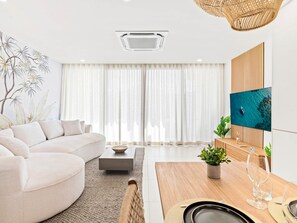A modern living room with a curved sofa, a flat-screen TV, and a dining area with a wooden table and chairs.