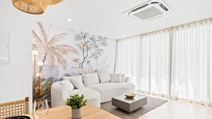 Ein modernes Wohnzimmer mit Sofa, Couchtisch, Esstisch mit Stühlen, Wand-Klimaanlage und großem Fenster mit Vorhängen.