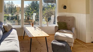 Een gezellige kamer met houten vloer, een bank, een stoel en een tafel. Grote ramen bieden uitzicht op bomen en een tuin.