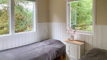 Una habitación pequeña y acogedora con dos camas, un aparador blanco y amplios ventanales con vistas a árboles.