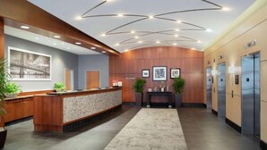 A modern reception area with a wooden reception desk, a large framed cityscape picture, and a console table with framed pictures. There are two elevators and a carpeted floor. - Near Independence Hall + Free Breakfast. Bar. Gym. (Philadelphia)