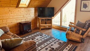 A cozy cabin interior with a wooden interior, a fireplace, a television, and a large window providing natural light. - Peaceful Rustic Mountain Getaway-Aspen Peaks Retreat (Hartsel)
