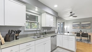 A modern kitchen with white cabinets, a stainless steel sink, and a dishwasher. There is a dining area with a table and chairs. - Cozy 5BD/fun house (Austin)