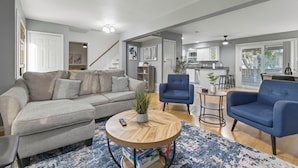 A modern living room with a grey sofa, blue armchairs, a wooden coffee table, and a kitchen in the background. - Cozy 5BD/fun house (Austin)