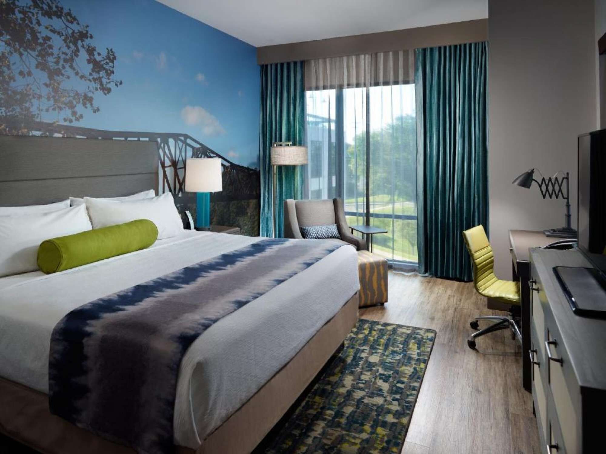 Ein Hotelzimmer mit einem großen Bett, einem Schreibtisch, einem Stuhl, einem Fernseher und einem Vorhangfenster.