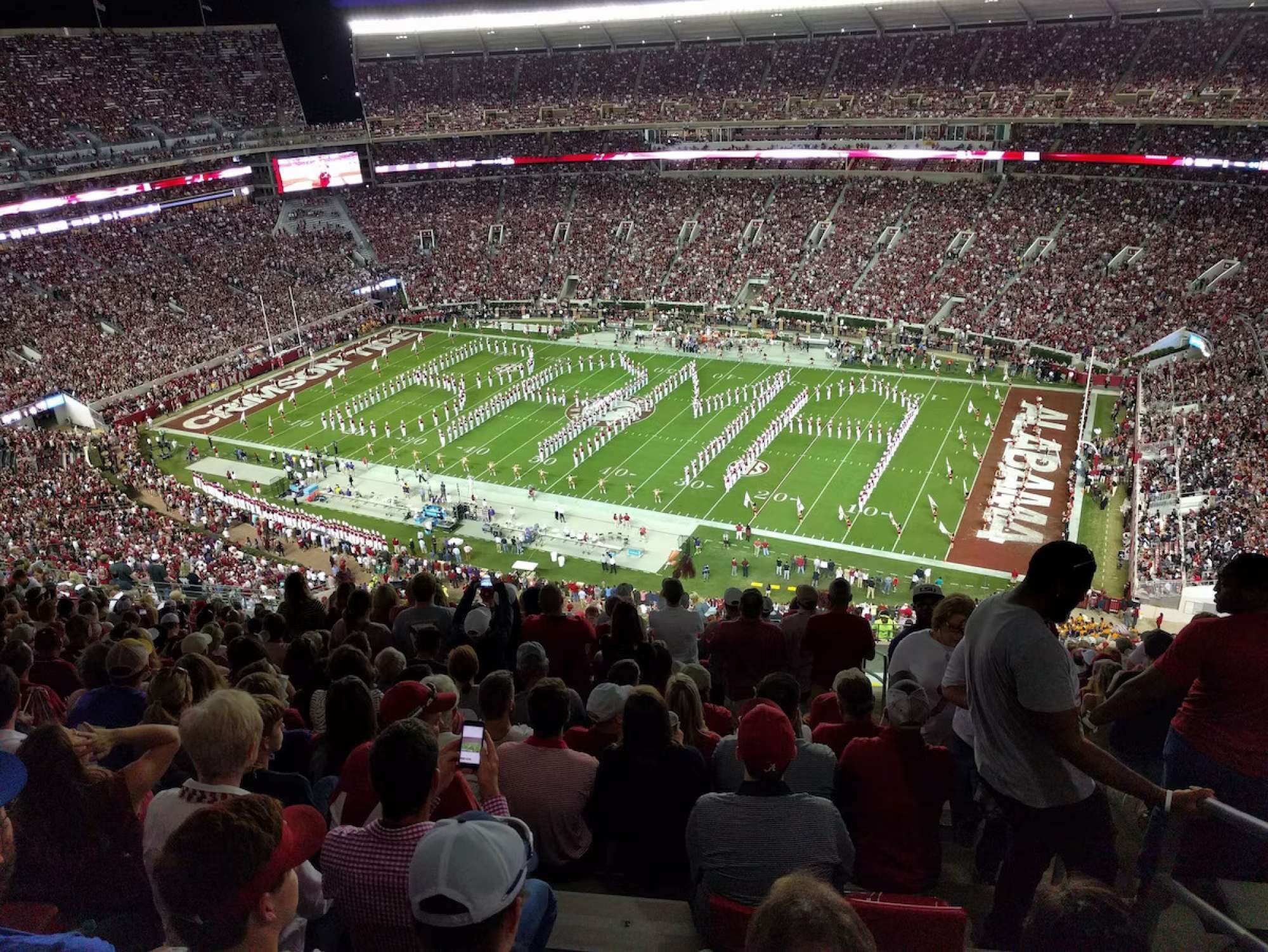 A stadium filled with spectators, featuring a football field with marching band formations.