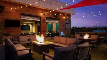 Uma área de estar ao ar livre com lareiras, um restaurante e vista para o horizonte da cidade ao entardecer.