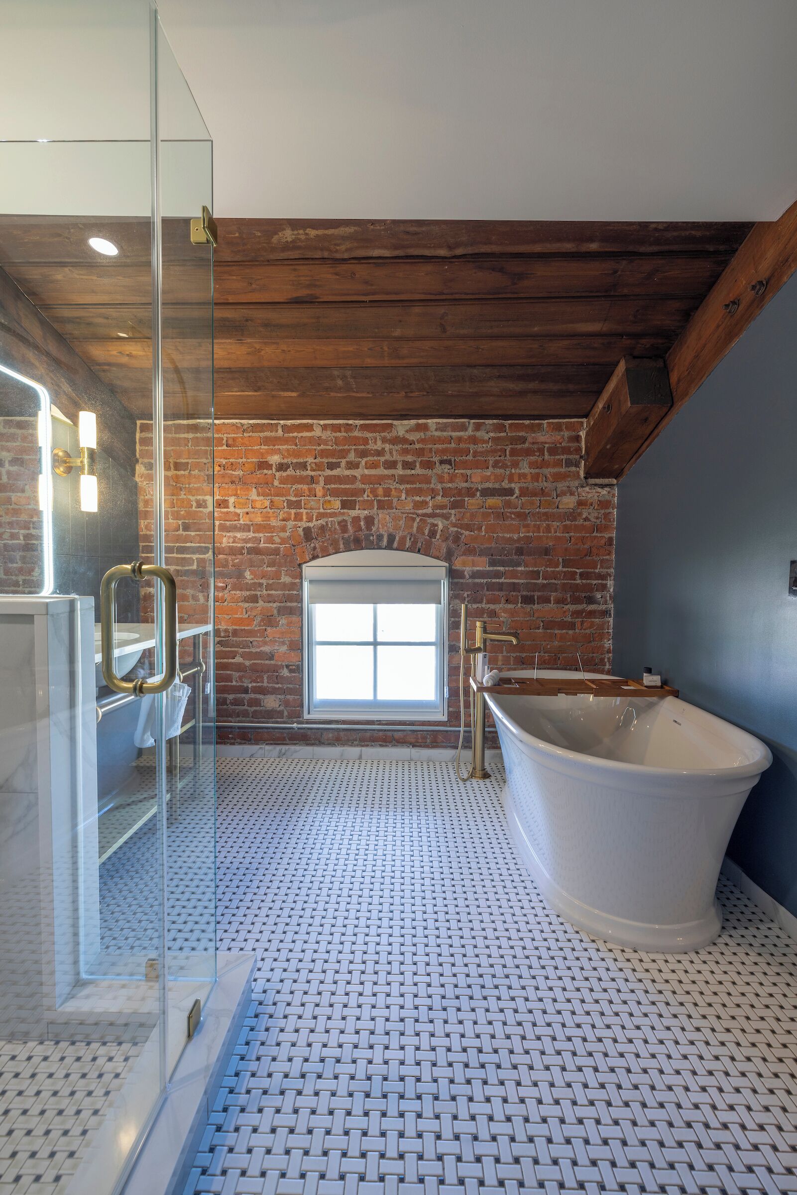 A bathroom with a glass shower enclosure, a freestanding bathtub, a brick wall, and a skylight.
