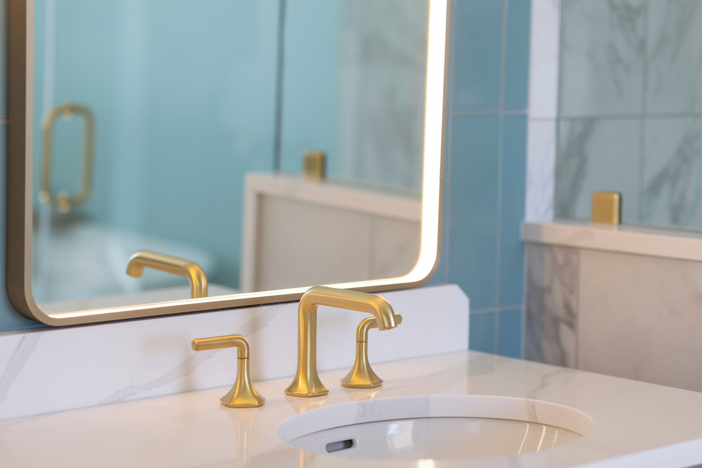 Ein Badezimmer mit einem weißen Marmorwaschbecken, goldenen Wasserhähnen und einem großen Spiegel, der einen Wasserhahn in Gold reflektiert.