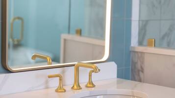 Ein Badezimmer mit einem weißen Marmorwaschbecken, goldenen Wasserhähnen und einem großen Spiegel, der einen Wasserhahn in Gold reflektiert.