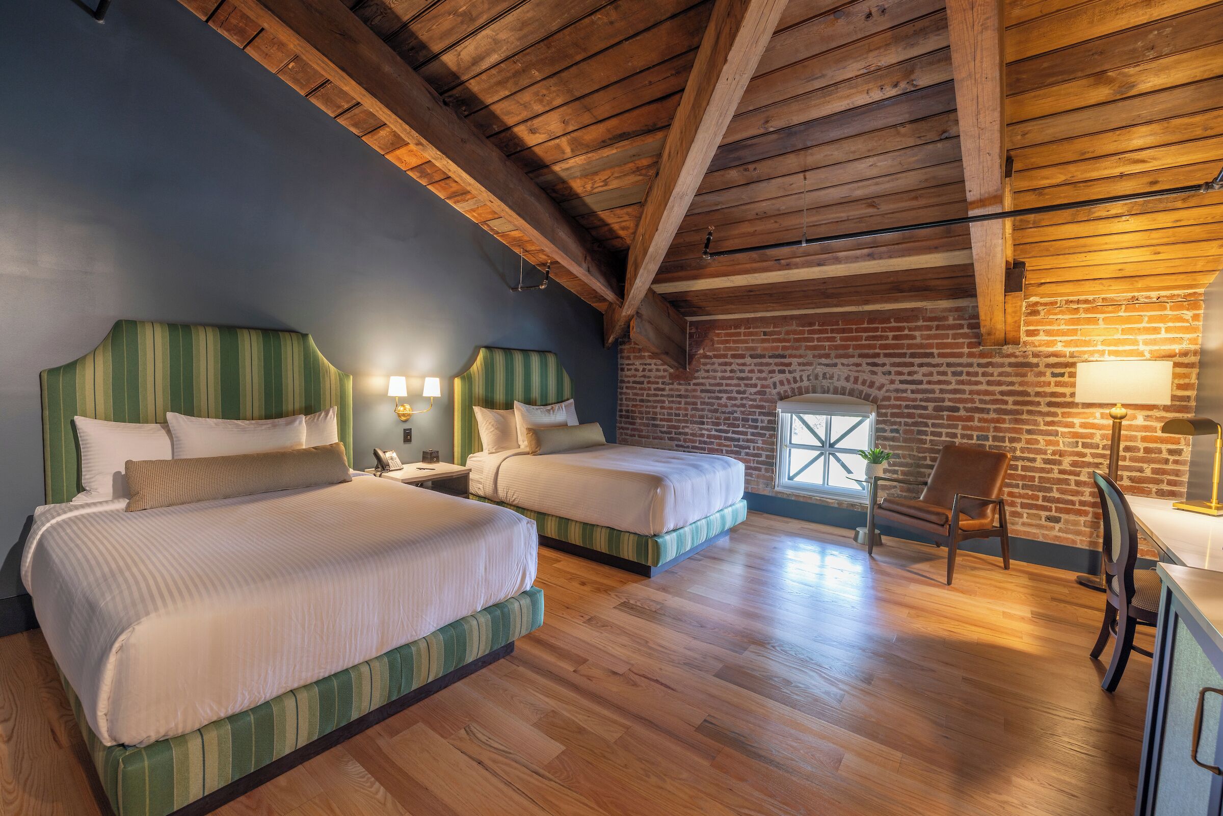 A room with two beds, wooden floors, a brick wall, and a window.