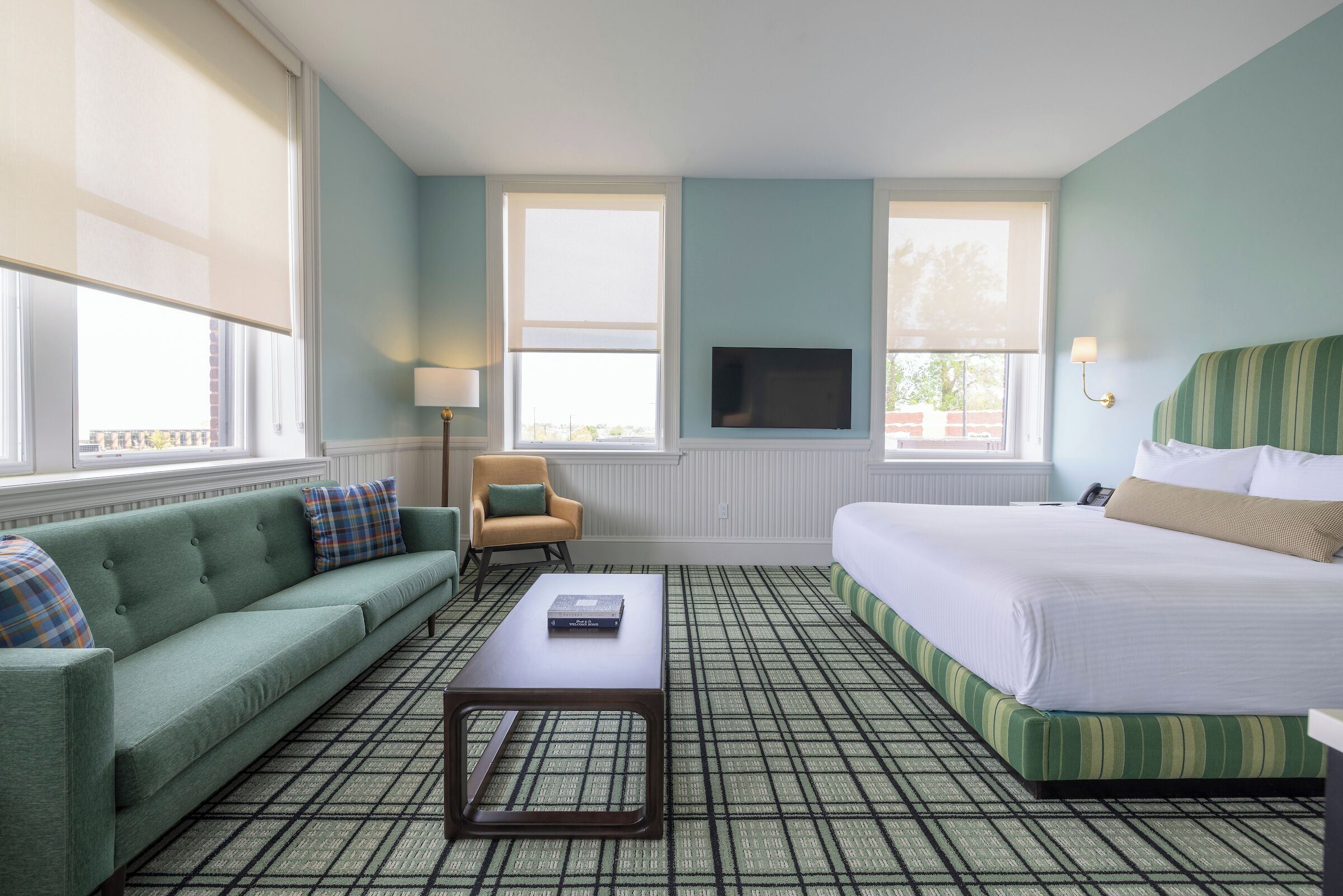 A hotel room with a green sofa, a bed with striped bedding, a television mounted on the wall, and a patterned carpet.