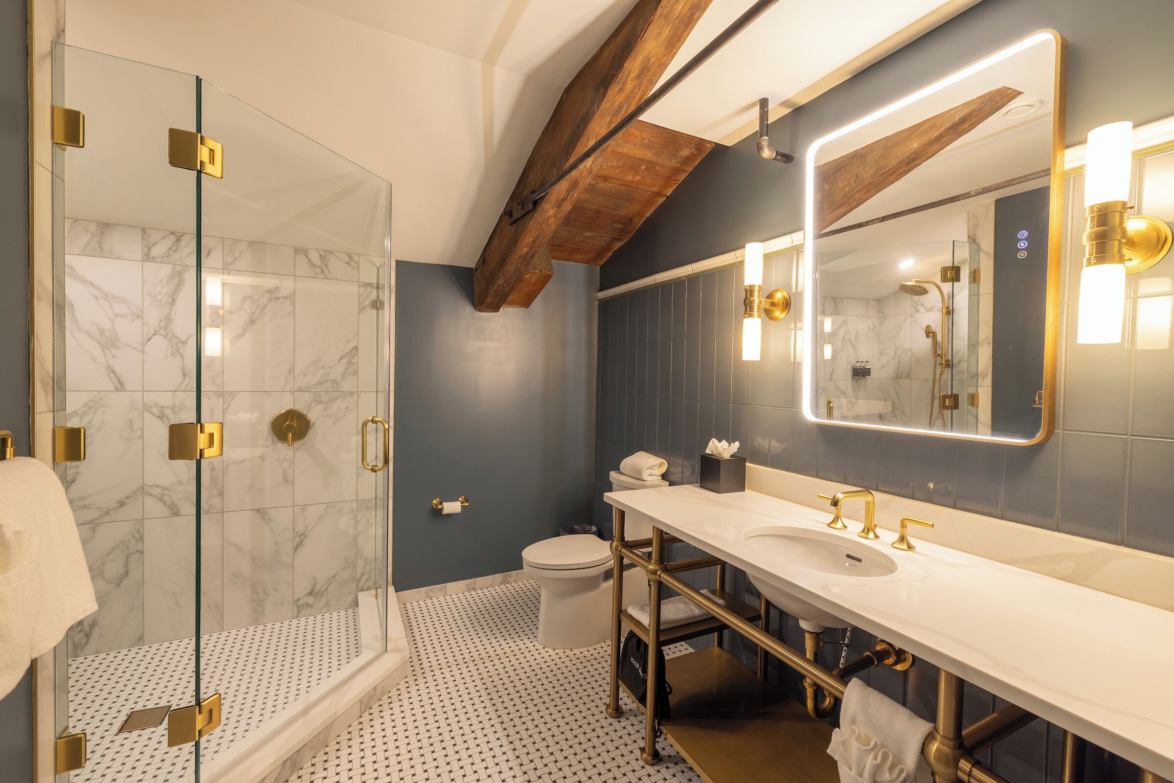 Ein modernes Badezimmer mit Duschkabine aus Glas, Waschbecken in Weiß mit goldenen Armaturen und einem großen Spiegel mit integrierter Beleuchtung.