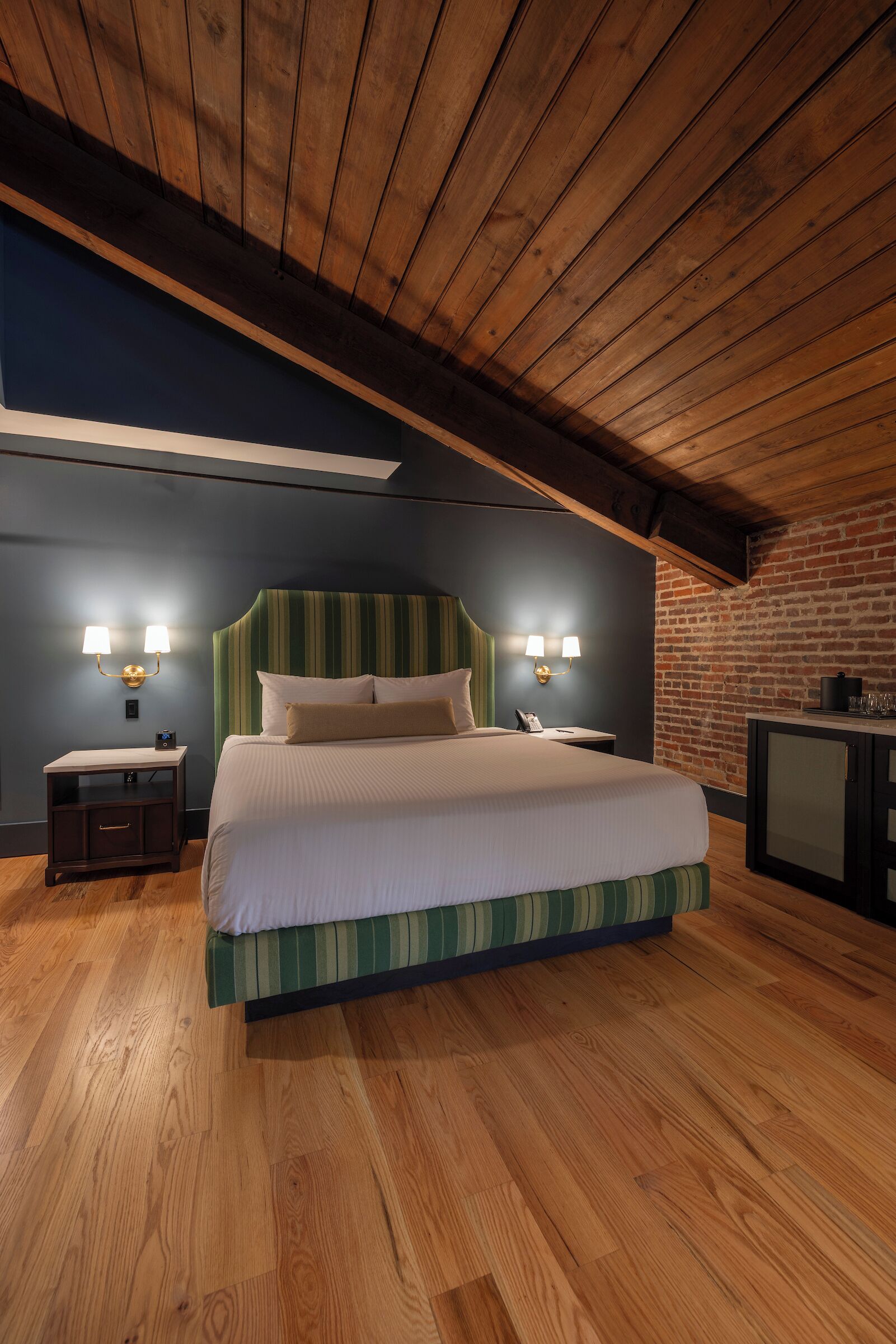 Een slaapkamer met een houten plafond, een bakstenen muur, een bed met gestreepte beddengoed, twee nachtlampjes, een nachtkastje en een klein kastje.