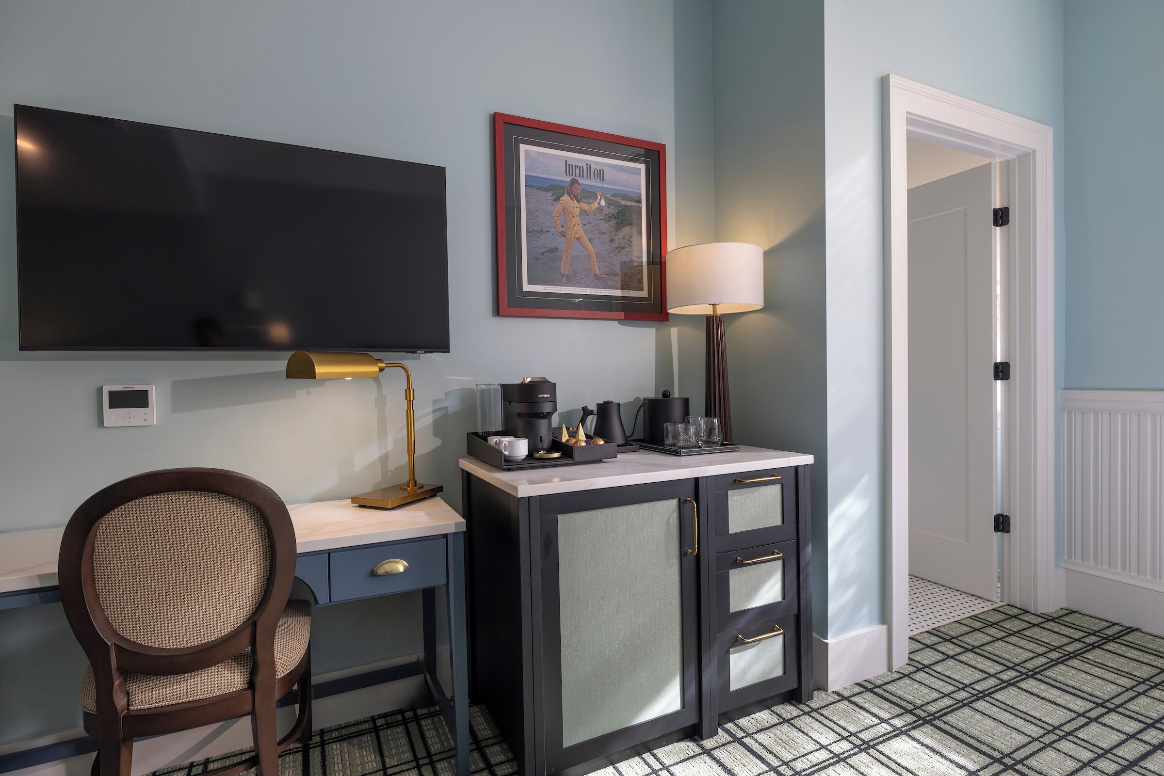 A room with a desk, chair, television, framed picture, lamp, and a door.
