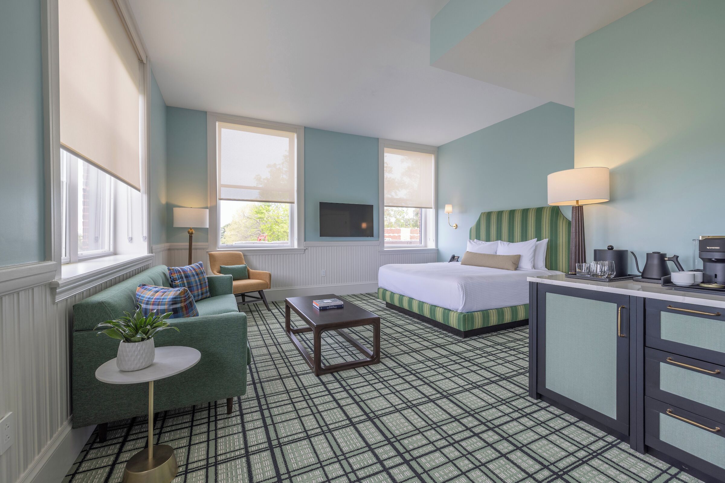 A hotel room with a green sofa, a bed, a small table, a kitchenette, and a TV.