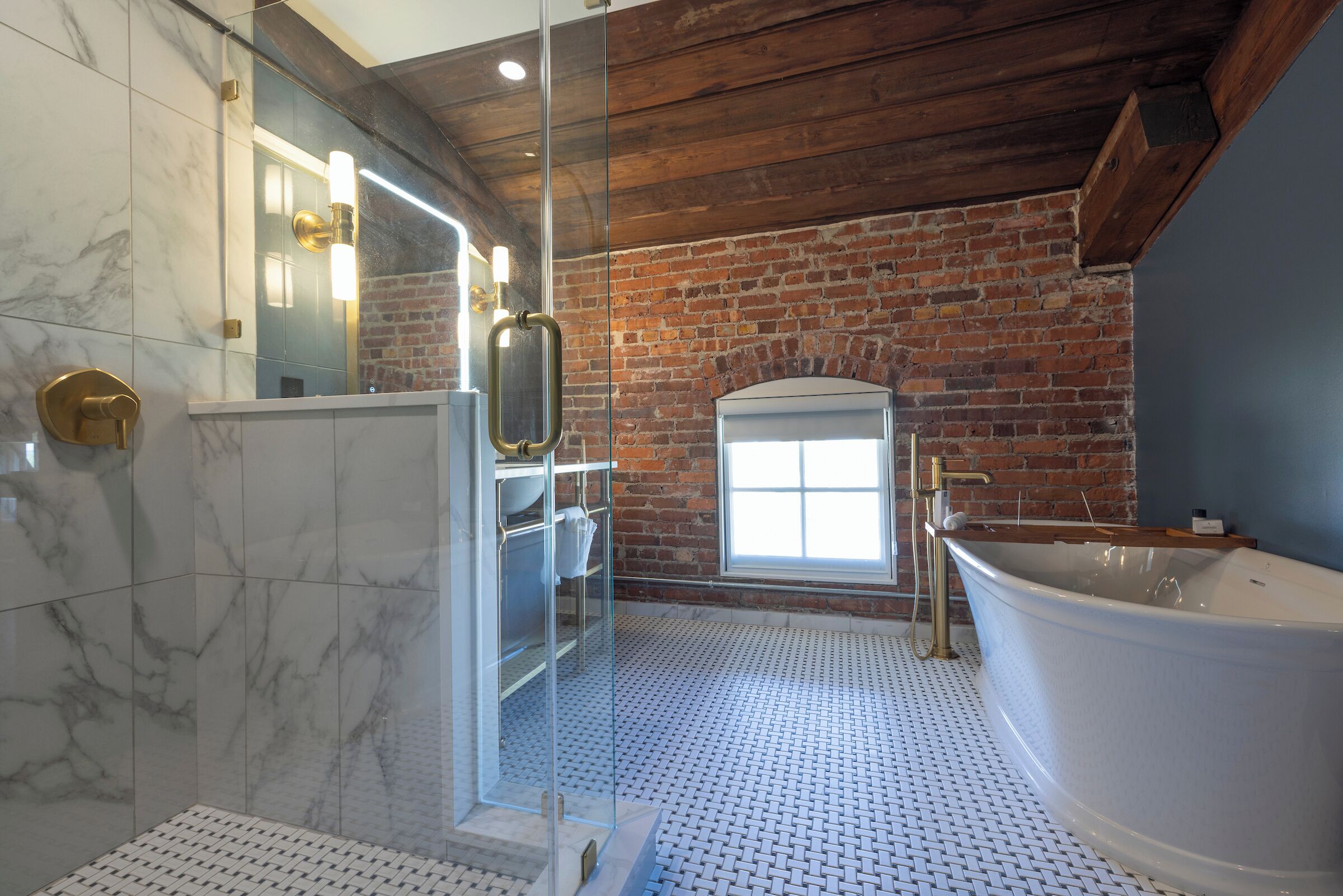 A bathroom with a large bathtub, a glass shower enclosure, a brick wall, and a window.