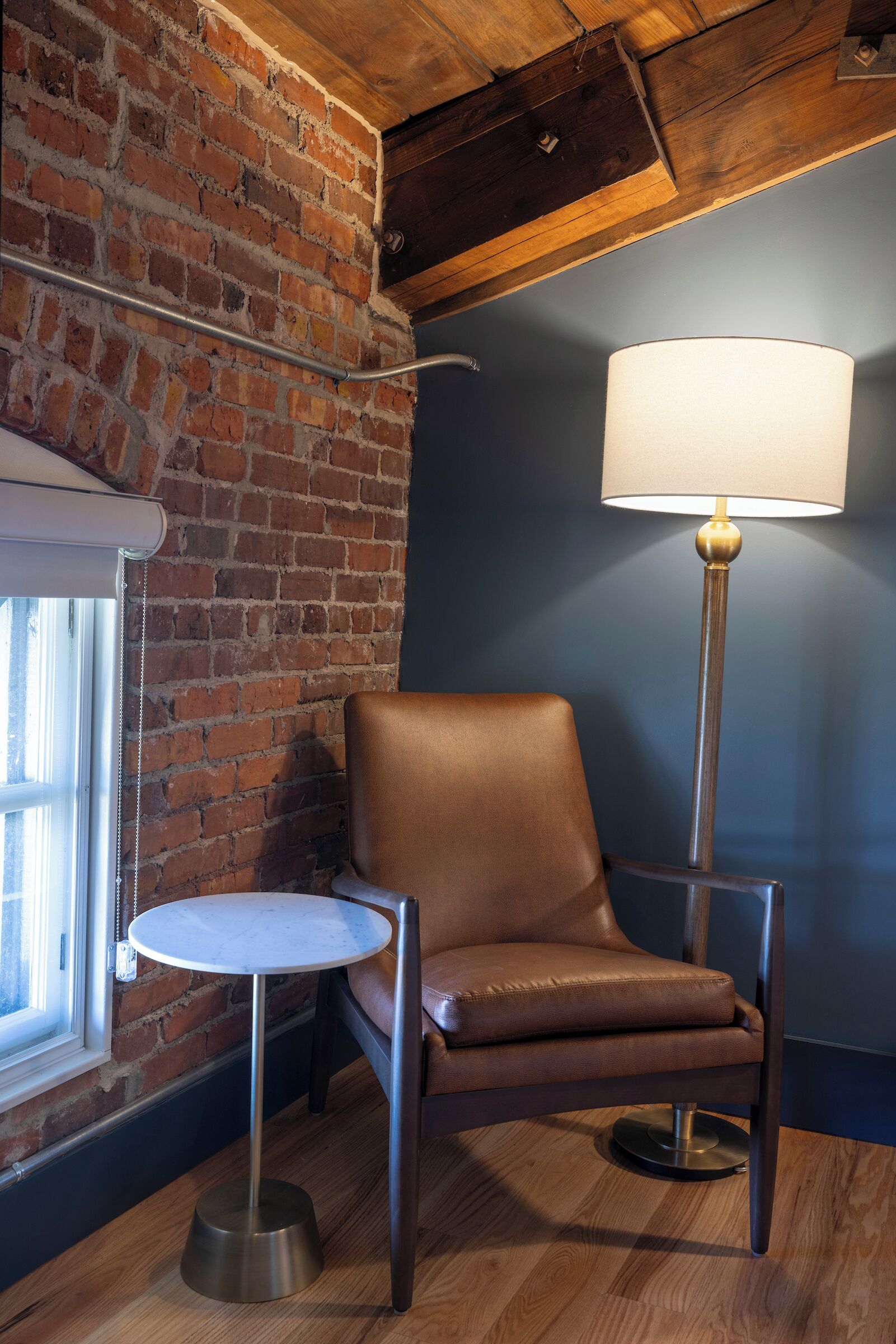 A room with a brick wall, a brown leather chair, a small round table, and a floor lamp.