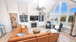 A spacious living room with a large brown leather sofa, a flat-screen TV mounted above a fireplace, and a ceiling fan. - Semi-Oceanfront, Ocean Views, Priv. Pool, Hot Tub-WL922, Beauty and the Beach (Corolla)