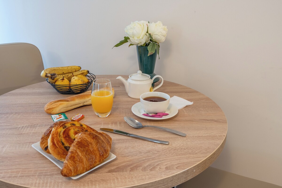 A breakfast table with croissants, a fruit basket, a teapot, a cup of coffee, and a vase of flowers.