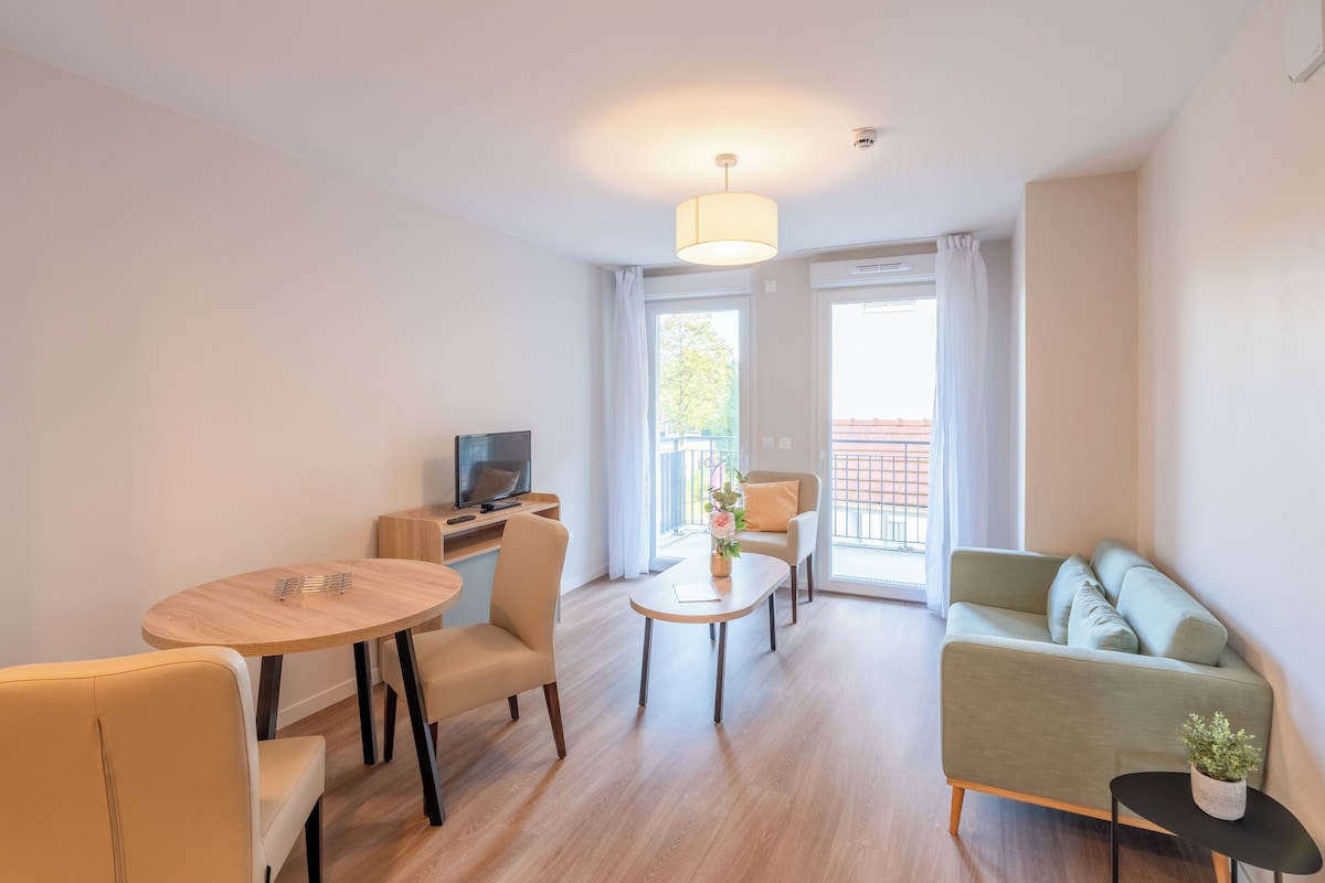 A modern living room with a sofa, a dining table, chairs, a TV, and a balcony with curtains.