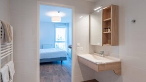 Un baño moderno con tocador de madera, un espejo grande y vista a un dormitorio con cama y ventana.