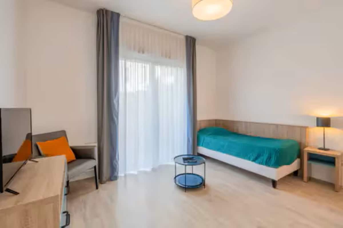 Habitació d'hotel amb un llit, una cadira, una taula petita, una làmpada i una finestra amb cortines.