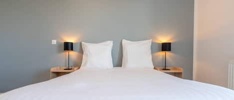 Una cama bien tendida con sábanas blancas, dos mesitas de noche con lámparas negras, y una pared de color gris claro.