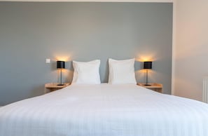 Una cama bien tendida con sábanas blancas, dos mesitas de noche con lámparas negras, y una pared de color gris claro.