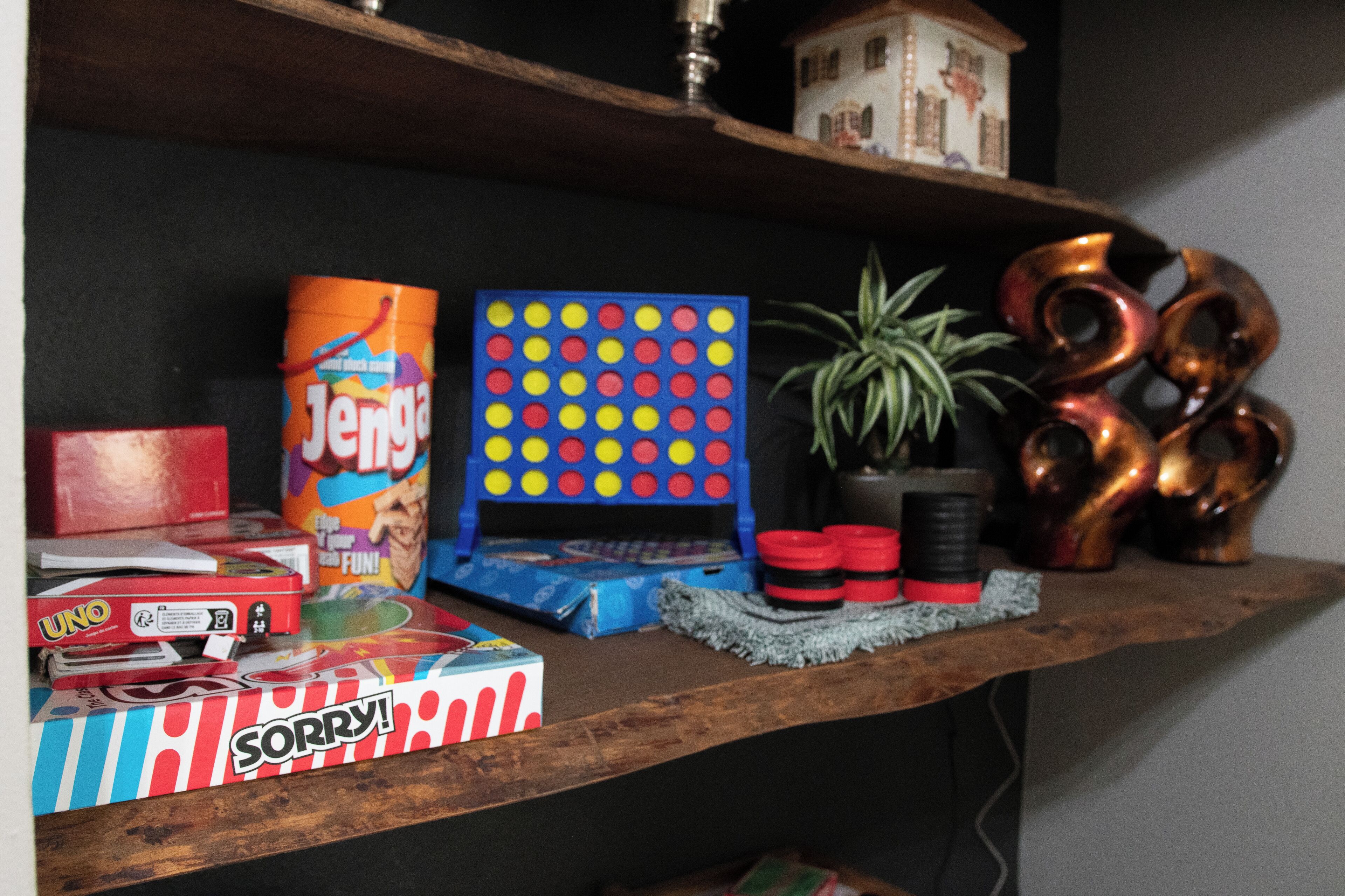 Un estante con varios juegos de mesa, incluyendo Jenga, UNO y Sorry!, además de elementos decorativos como una planta y esculturas de metal.