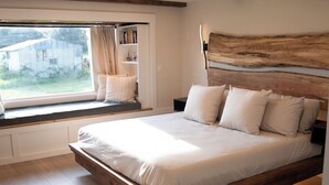 A bedroom with a large bed, a window seat with books, and a view of a house.