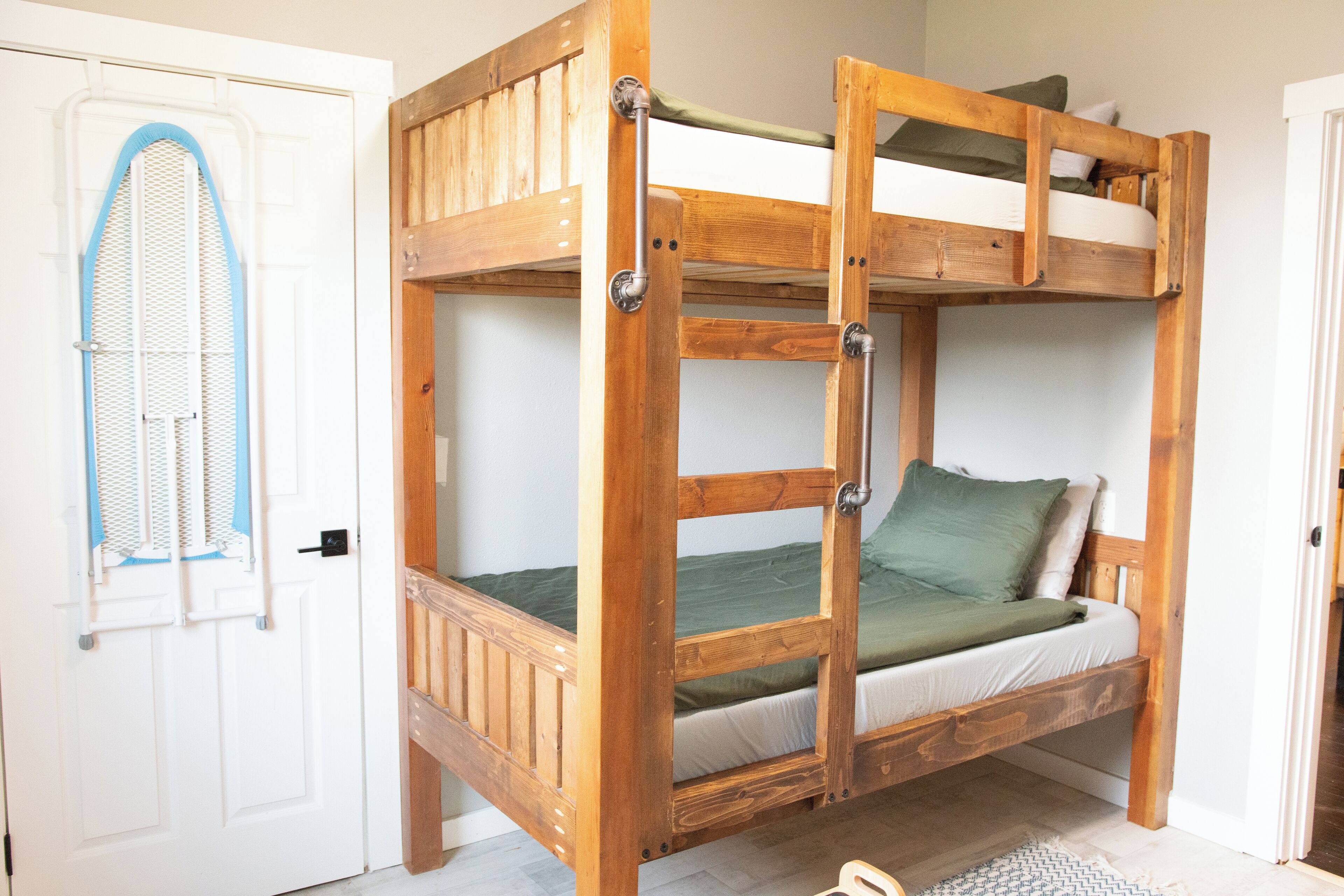 Una cama litera con estructura de madera, colchón verde y escalera metálica.