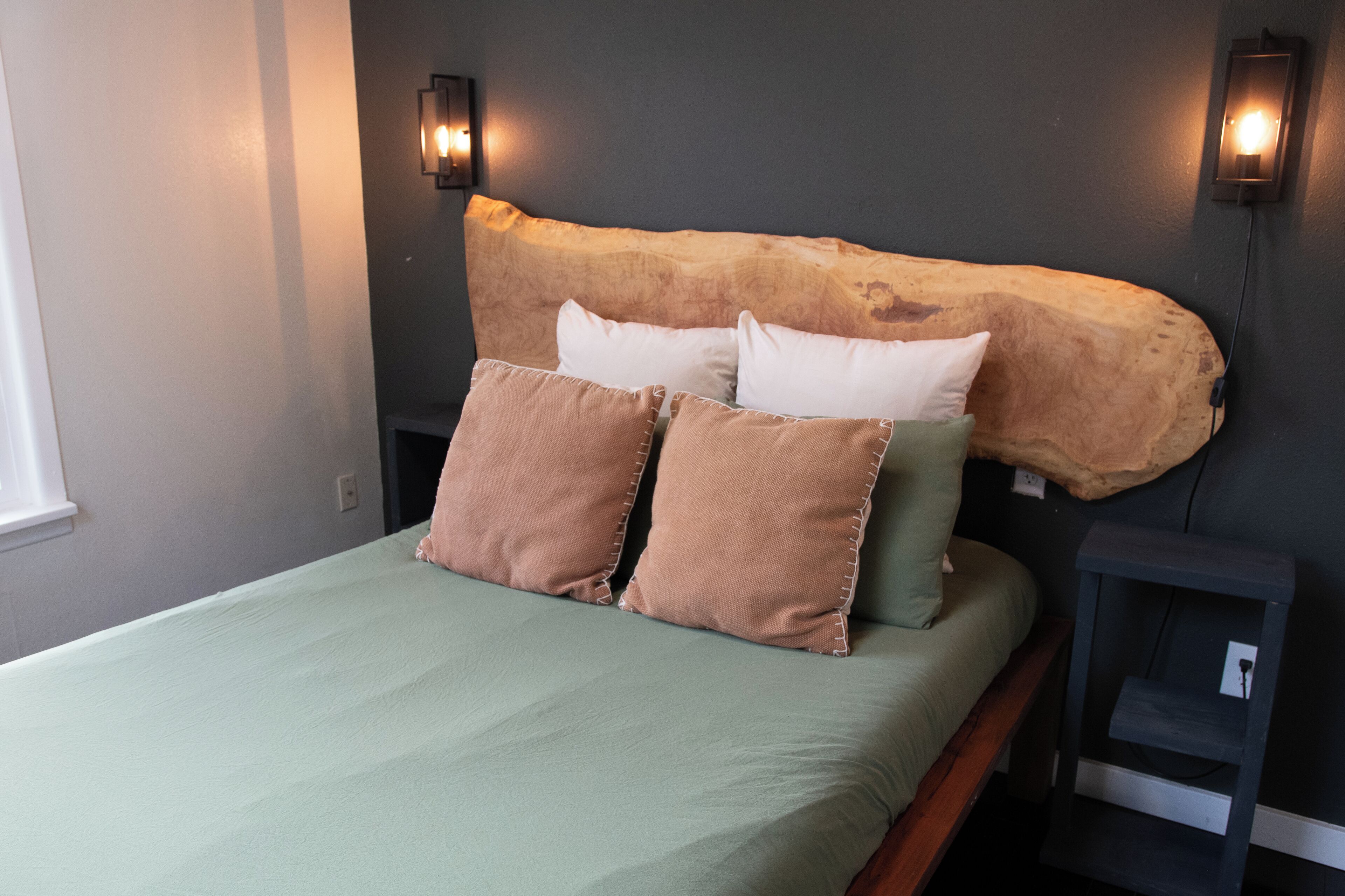 A bed with a green bedspread, two pillows, and a headboard made of a natural material. There are two wall-mounted light fixtures on either side of the headboard.