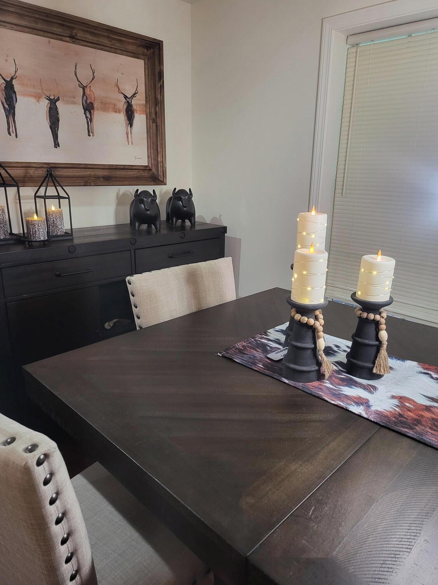 A dining table with a dark wood finish, a framed artwork above it, and a window with blinds to the right.