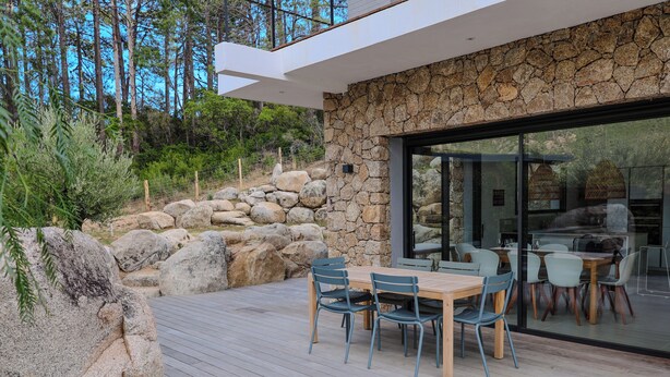 Uma área moderna de refeições ao ar livre com um muro de pedra e um deck de madeira, com vista para uma encosta rochosa e uma floresta.
