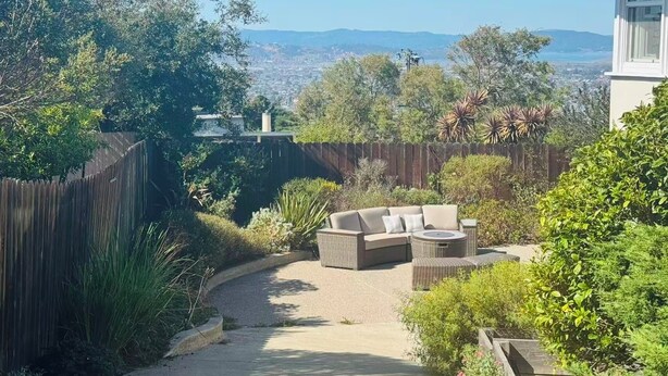 Un patio amueblado al aire libre, una valla de madera y vistas a un paisaje urbano y montañas distantes.