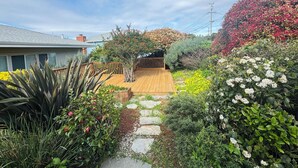 Un jardín con terraza de madera, sendero de piedra y diversas plantas y flores.