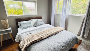 Un dormitorio con cama, mesita de noche y dos ventanas con cortinas.