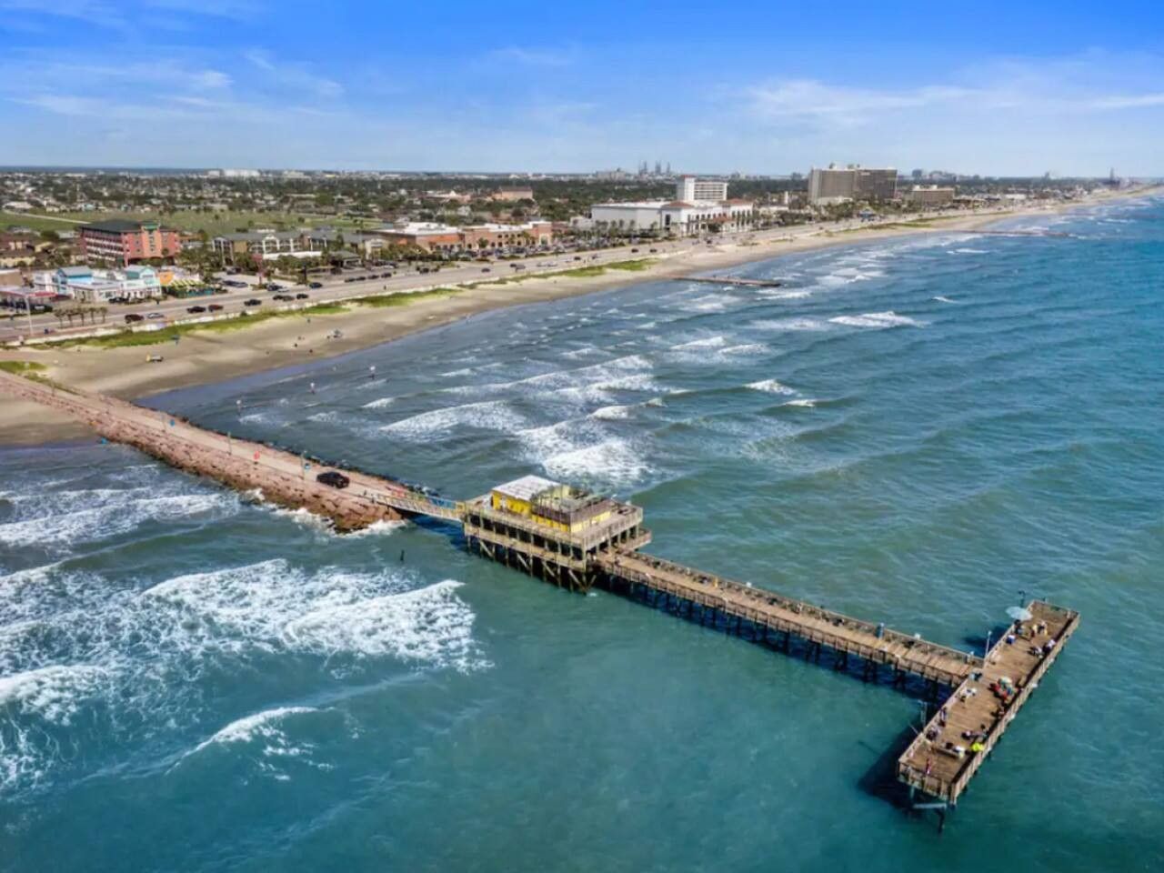 Eine Pier, die sich ins Meer erstreckt, mit einem Gebäude darauf, einem Strand und einer Stadtlandschaft im Hintergrund.